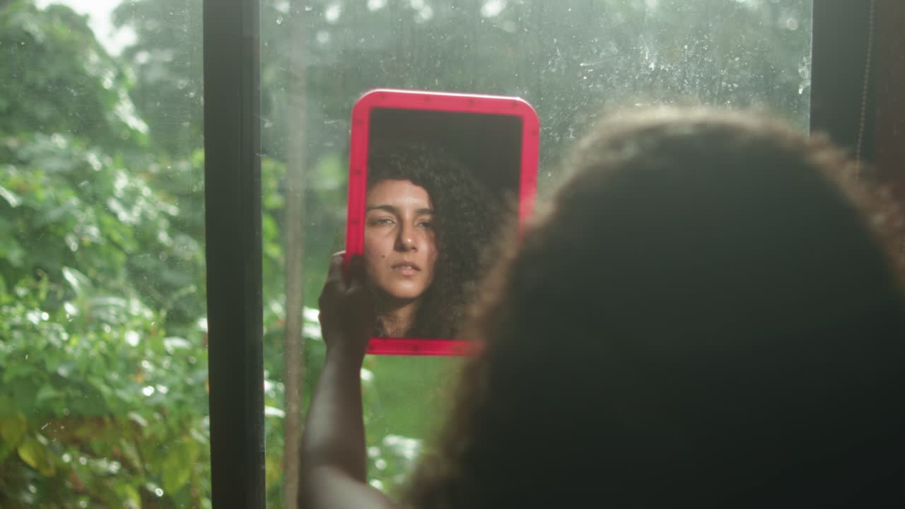 Woman with curly hair holding mirror indoors near window