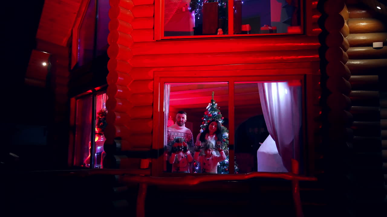 Happy young couple and kids waiting for Santa at home. Family celebrating Christmas in cosy house. Family members standing near window and looking for Christmas miracle. View from outdoors.