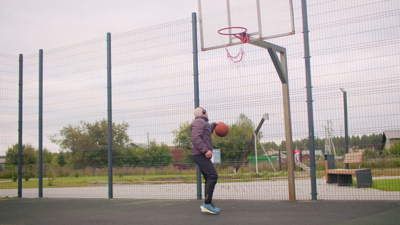 Uomo che si esercita nei tiri a canestro all'aperto su un campo urbano, indossa una felpa con cappuccio e un berretto, si concentra su esercizi di layup e palleggio sotto un cielo nuvoloso, sfondo di un parco vuoto con recinzione a catena e tabellone