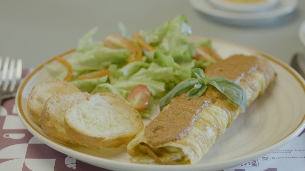 Close-up of an appetizing dish served with a rolled egg tortilla filled and covered with sauce, accompanied by a fresh vegetable salad and slices of bread