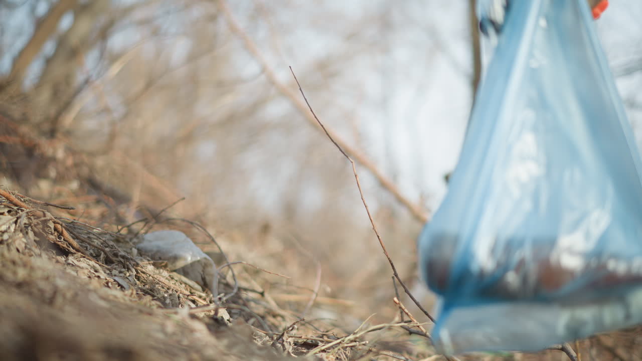 Person wearing orange gloves placing collected trash into blue plastic bag during outdoor cleanup, removing litter from dry natural area to support environmental protection, waste reduction