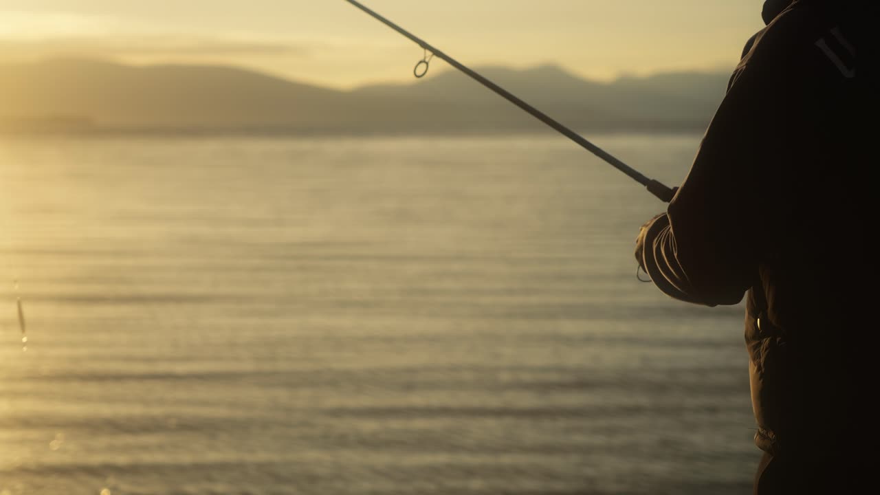Primo piano di un pescatore che recupera la lenza con un pilker e una canna da pesca in vista lungo la riva, mentre la luce dorata luccica sull'acqua calma e le montagne distanti creano uno sfondo sereno e cinematografico.