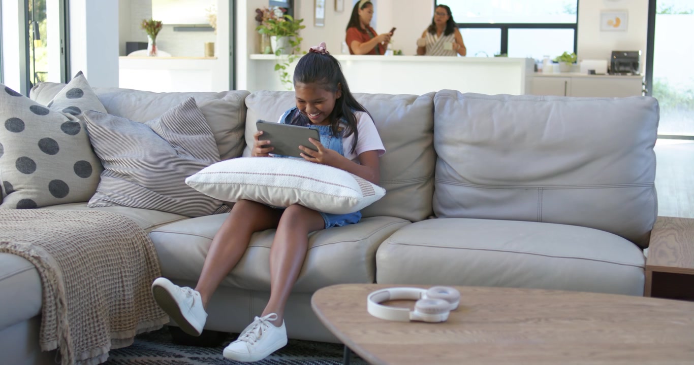 Ung kvinde fra en mangfoldig familie ser sjovt tabletklip, griner og retter på puden i sofaen.