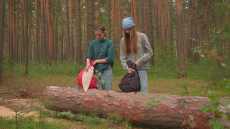 hikers approach fallen tree in forest, each holding backpacks, one with blue bandana struggles to unzip her bag, while the other places hers on the log, both focused on their task amidst tall trees