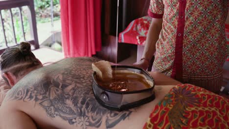 el terapeuta del spa vierte lentamente aceite tibio en el recipiente revestido de cera en la parte posterior de un paciente masculino que disfruta de un tratamiento kati vasti en un retiro ayurvédico