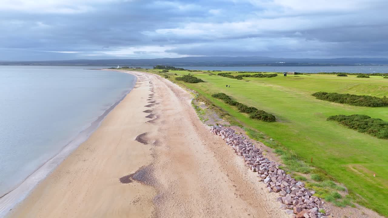 Drone footage glides above a sandy shoreline and adjacent golf course under overcast skies, capturing the tranquil Scottish Highlands coastline with smooth, steady movement