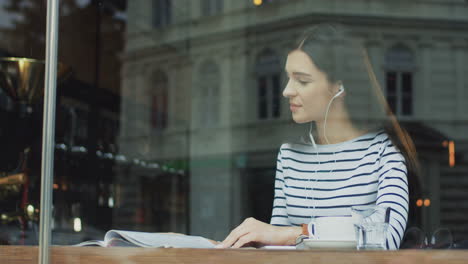 Mujer-Morena-Sentada-Detrás-De-La-Ventana-Del-Café,-Escuchando-Música-Y-Pasando-Las-Páginas-De-Una-Revista