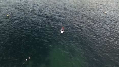 Man-kneeled-on-stand-up-paddle-paddling-close-to-yacht,-aerial-orbiting-overhead-view