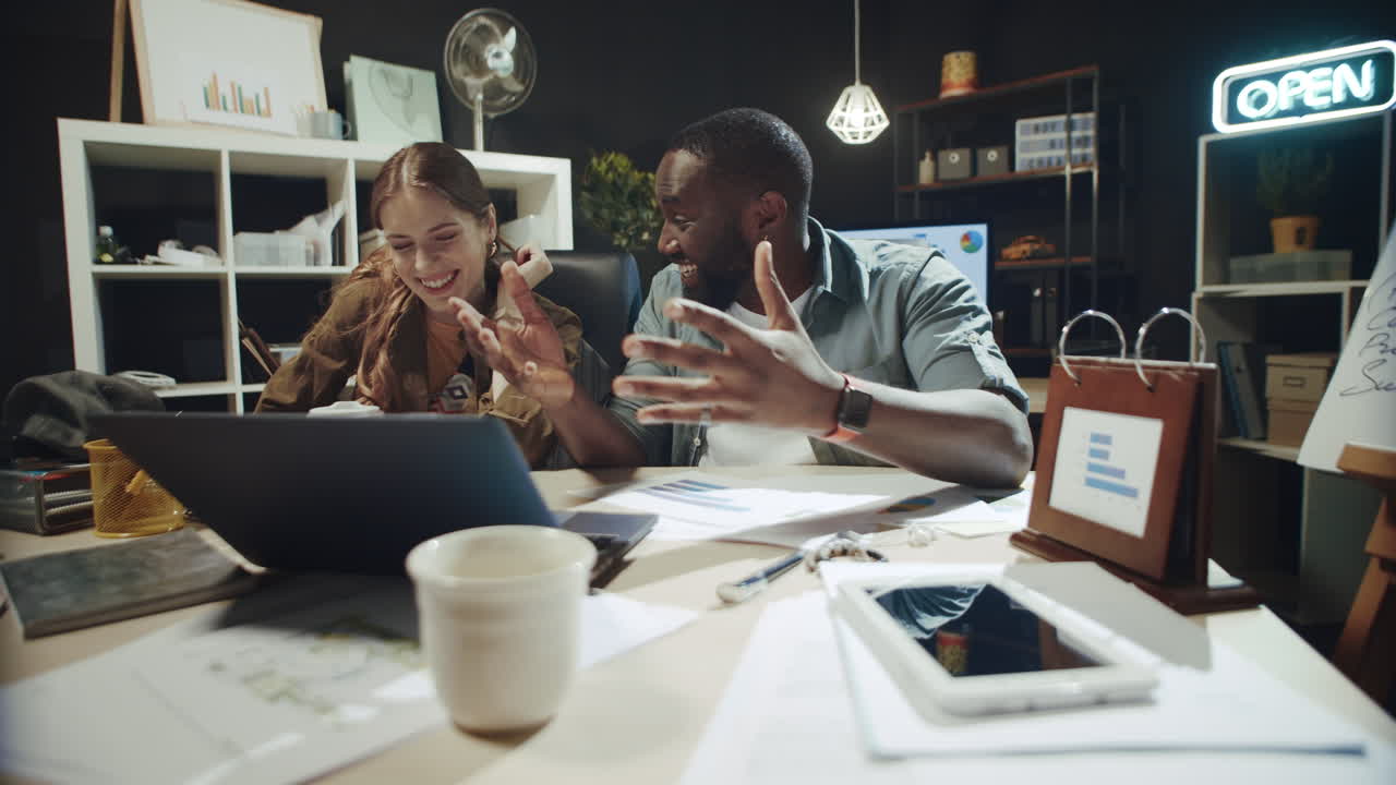 un hombre afro sonriente contando una historia a una mujer hipster cerca de una computadora portátil en una oficina oscura.