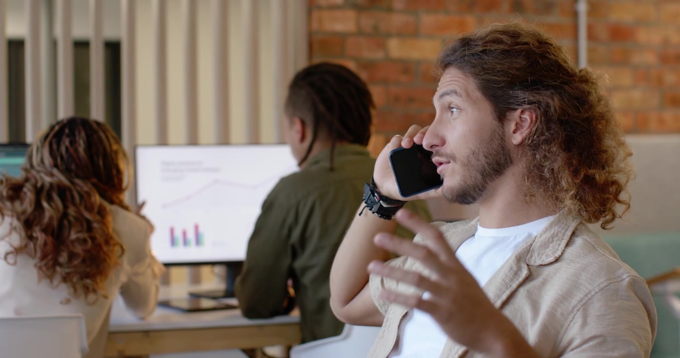 Diverse coworkers watching adult man on right taking call, gesturing, discussing charts at desk