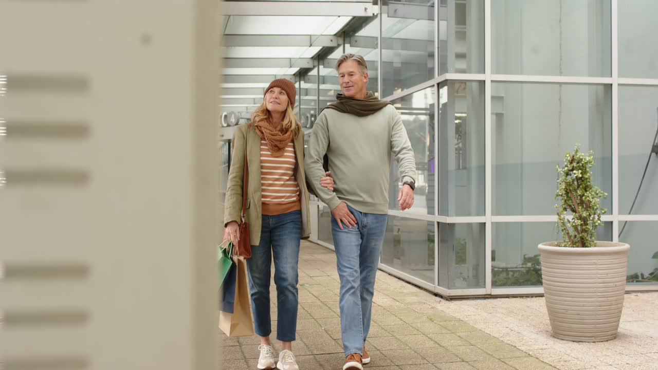Dopo aver fatto shopping, coppia di anziani che cammina a braccetto lungo il camminamento sotto la tettoia portando borse della spesa