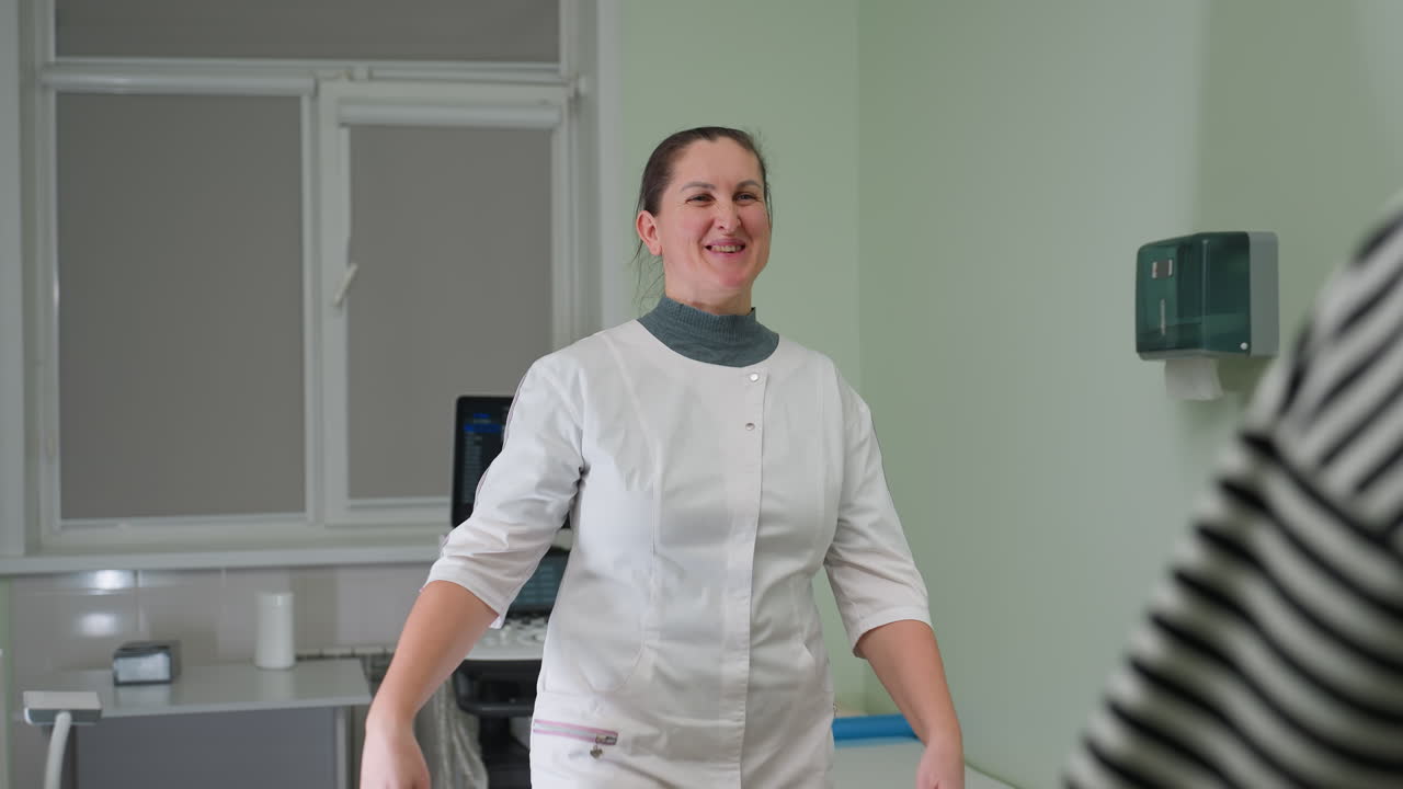 Female gynecologist stands in clinic room smiling warmly as she welcomes woman with affectionate hug while patient takes seat on medical bed, radiating calm atmosphere in professional environment