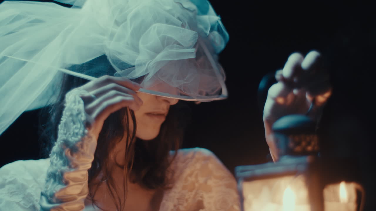 A woman in a white dress and veil holds a lit lantern in the dark