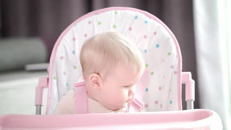 Mother-putting-baby-in-chair-in-kitchen.-Mom-hands-fasten-belts-in-pink-chair