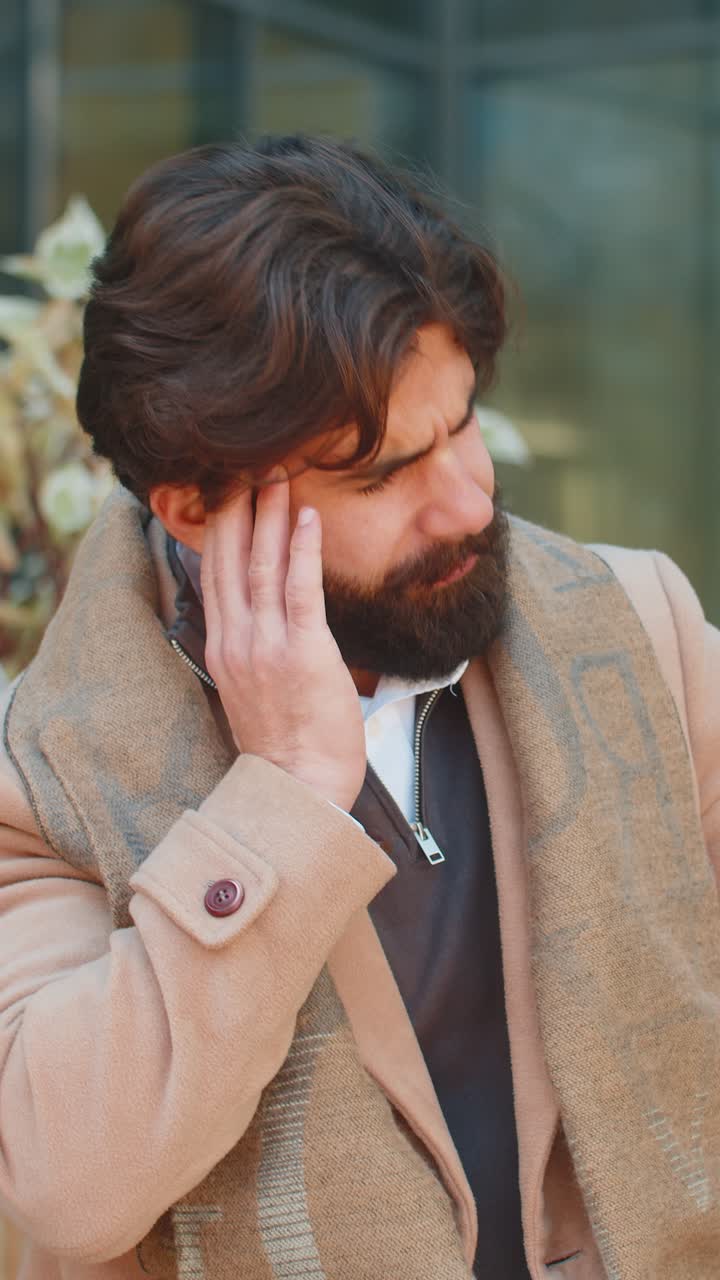 Sick young indian business man feels headache while sitting against building on city street
