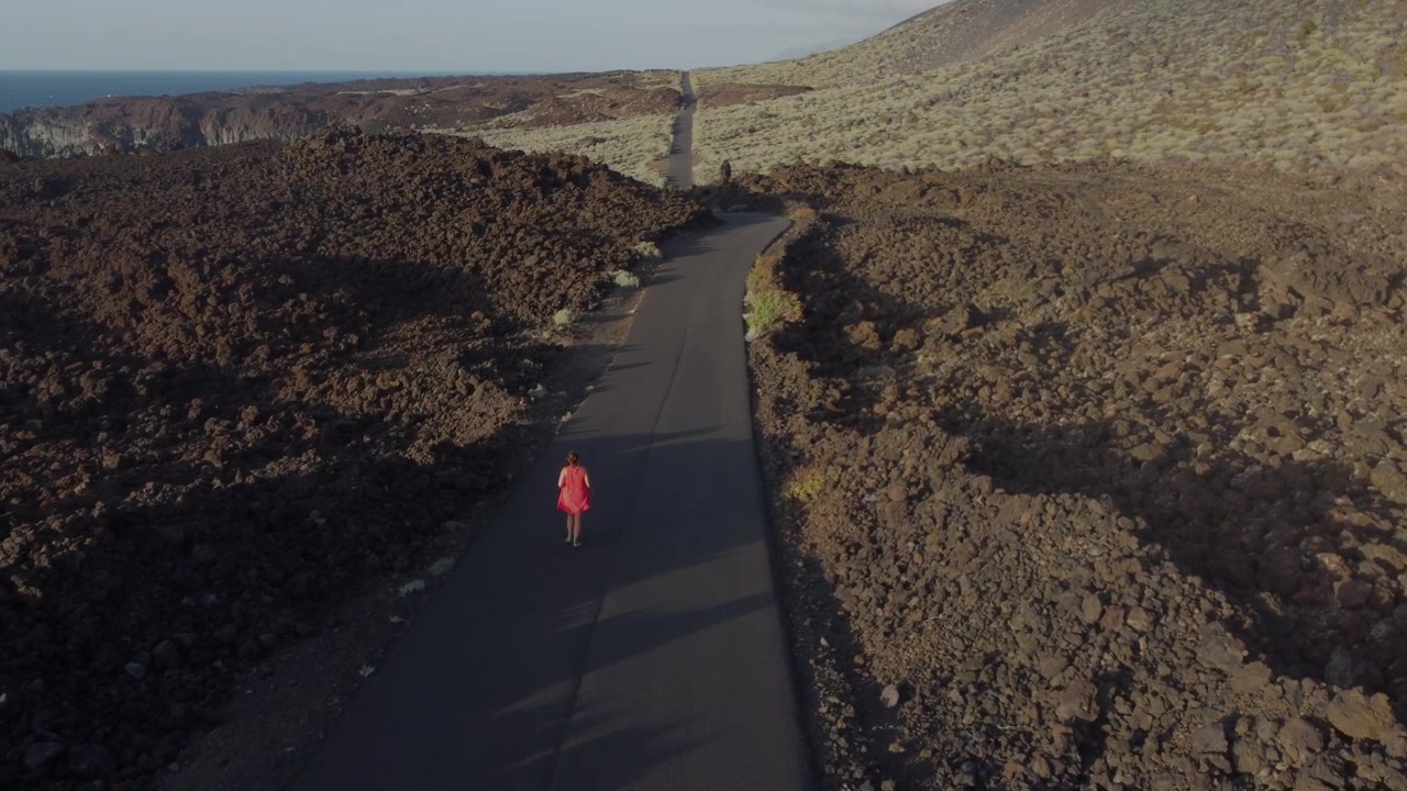 Una donna in abito rosso che sventola al vento corre lungo una strada dritta e deserta in un luogo dopo attività vulcanica - isola di El Hierro, Spagna.