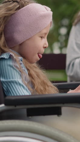 cute little girl in wheelchair takes funny selfies while mom reading news on smartphone. woman rests in park with daughter using gadgets closeup