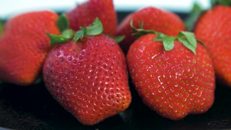 Fresh-big-red-tasty-ripe-strawberries-rotates-slowly-on-a-black-plate-on-light-blue-background,-healthy-food-concept,-extreme-close-up-shot,-camera-rotate-left