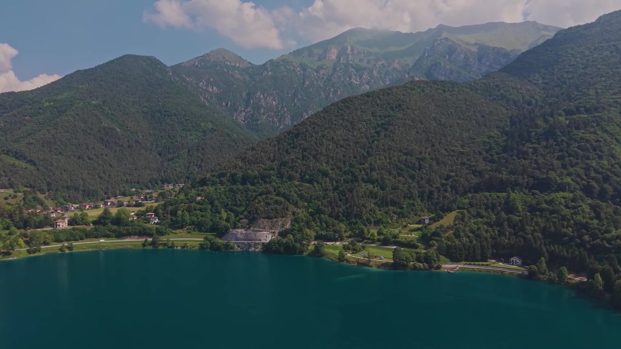 Turkuaz Ledro Gölü kıyı şeridinin, küçük bir alp köyü, göl kenarı yolu, yemyeşil ormanlık dağ yamaçları ve arka planda kayalık zirvelerle havadan drone görüntüsü, güneşli bir yaz günü.