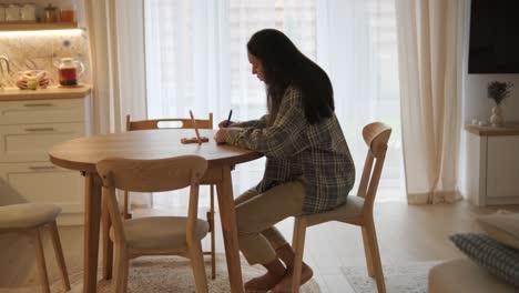 a pregnant woman sitting at a wooden table in a cozy, softly lit home interior, writing or journaling. the scene exudes calm, focus, and a sense of preparation during maternity