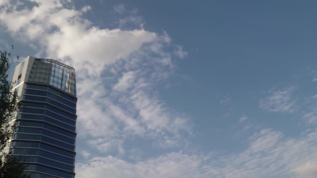 Uma filmagem time-lapse ampla em ângulo baixo captura nuvens suaves flutuando rapidamente através de um céu azul de verão acima de um moderno arranha-céu de escritórios de vidro no Infopark Budapeste, Hungria, em uma tarde ensolarada.
