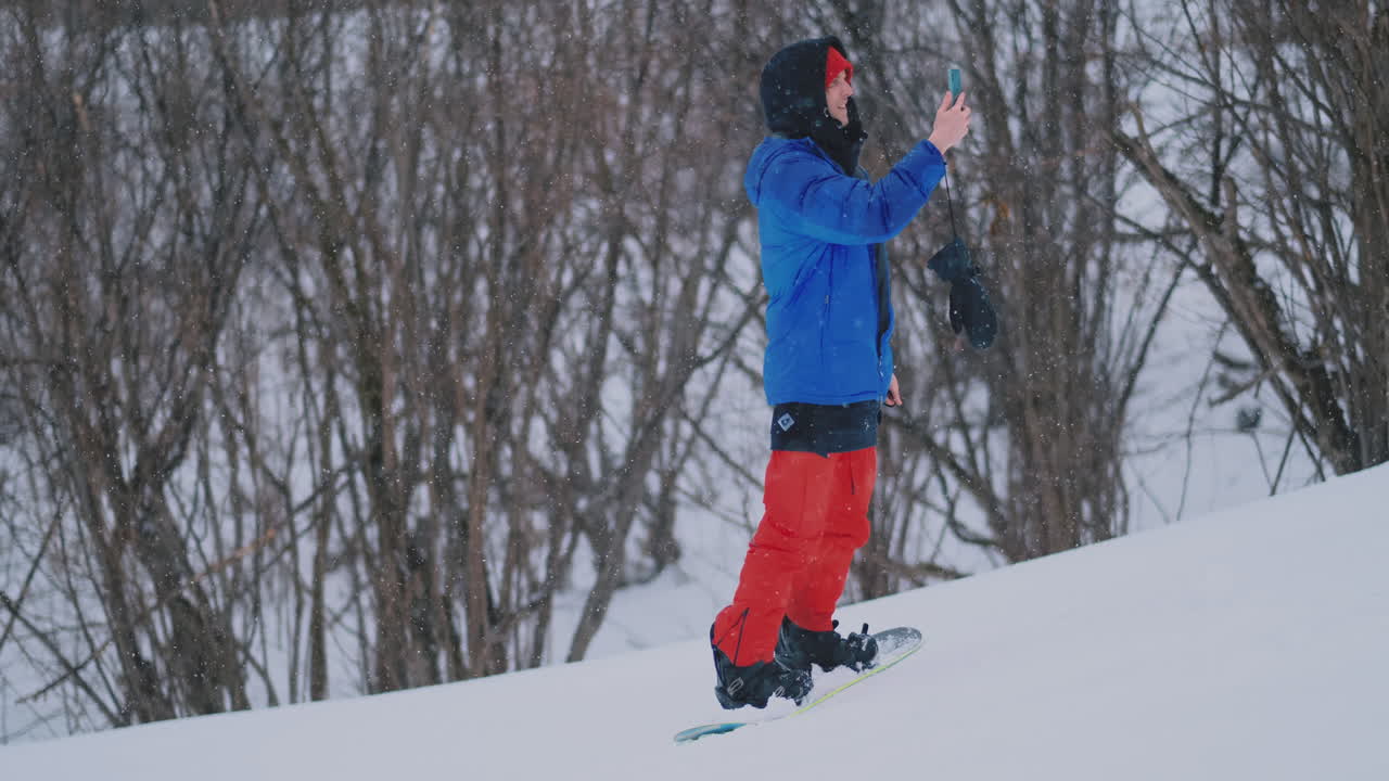 un snowboarder masculino monta la tabla en el esquí hasta la pista de nieve y escribe mensajes en el teléfono inteligente a sus amigos.