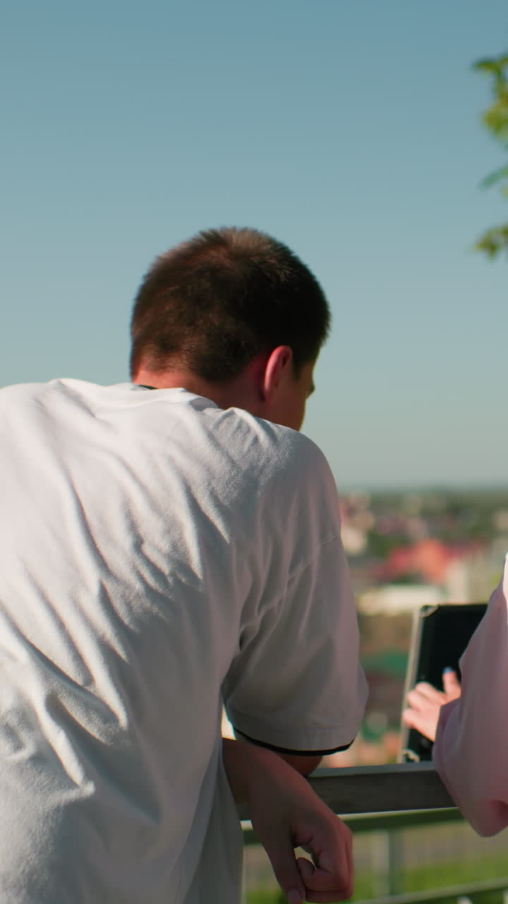 vista trasera de un niño y una niña inclinados sobre una barandilla de hierro mientras interactúan mientras la niña muestra algo en una tableta, el fondo presenta un paisaje urbano borroso y vegetación