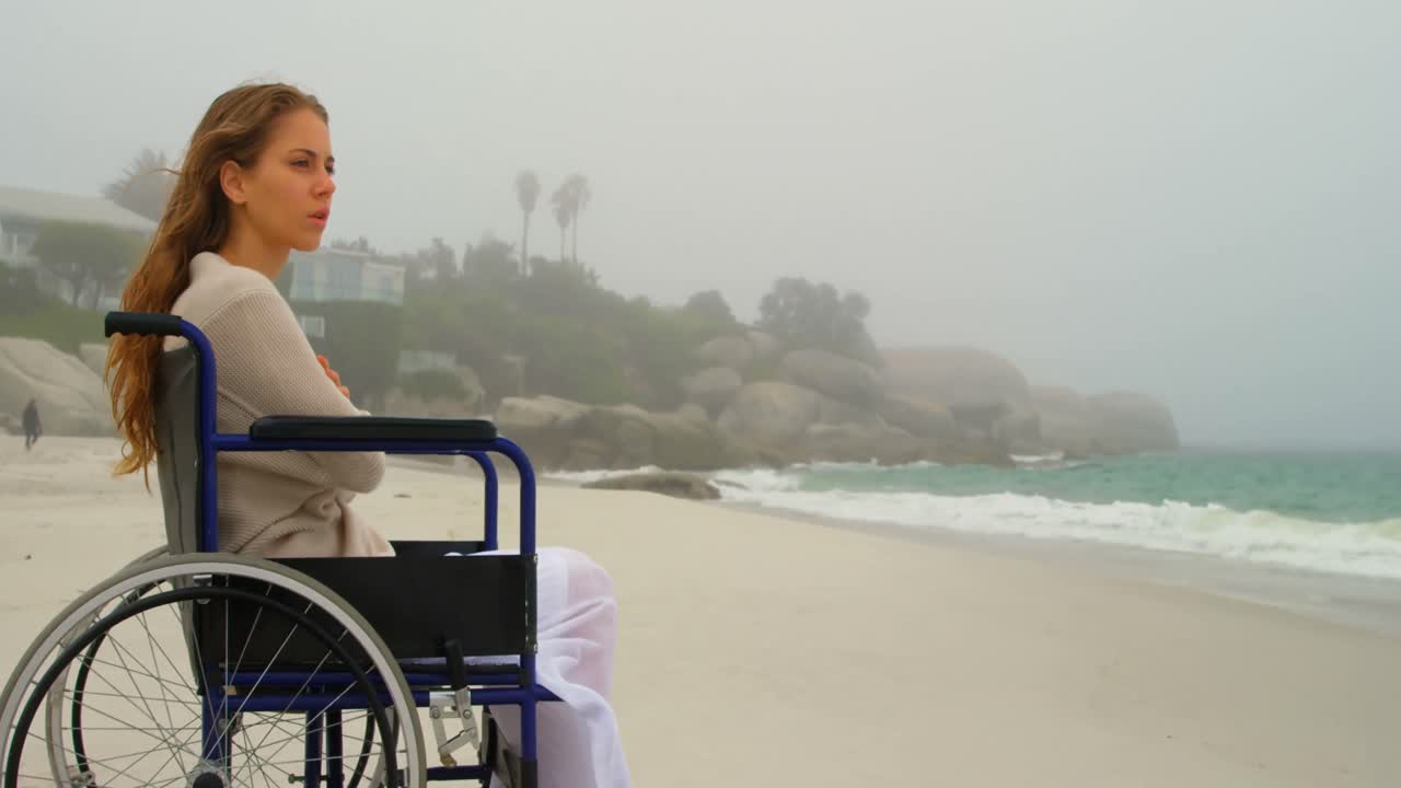 Side view of young Caucasian woman sitting on wheelchair at beach 4k