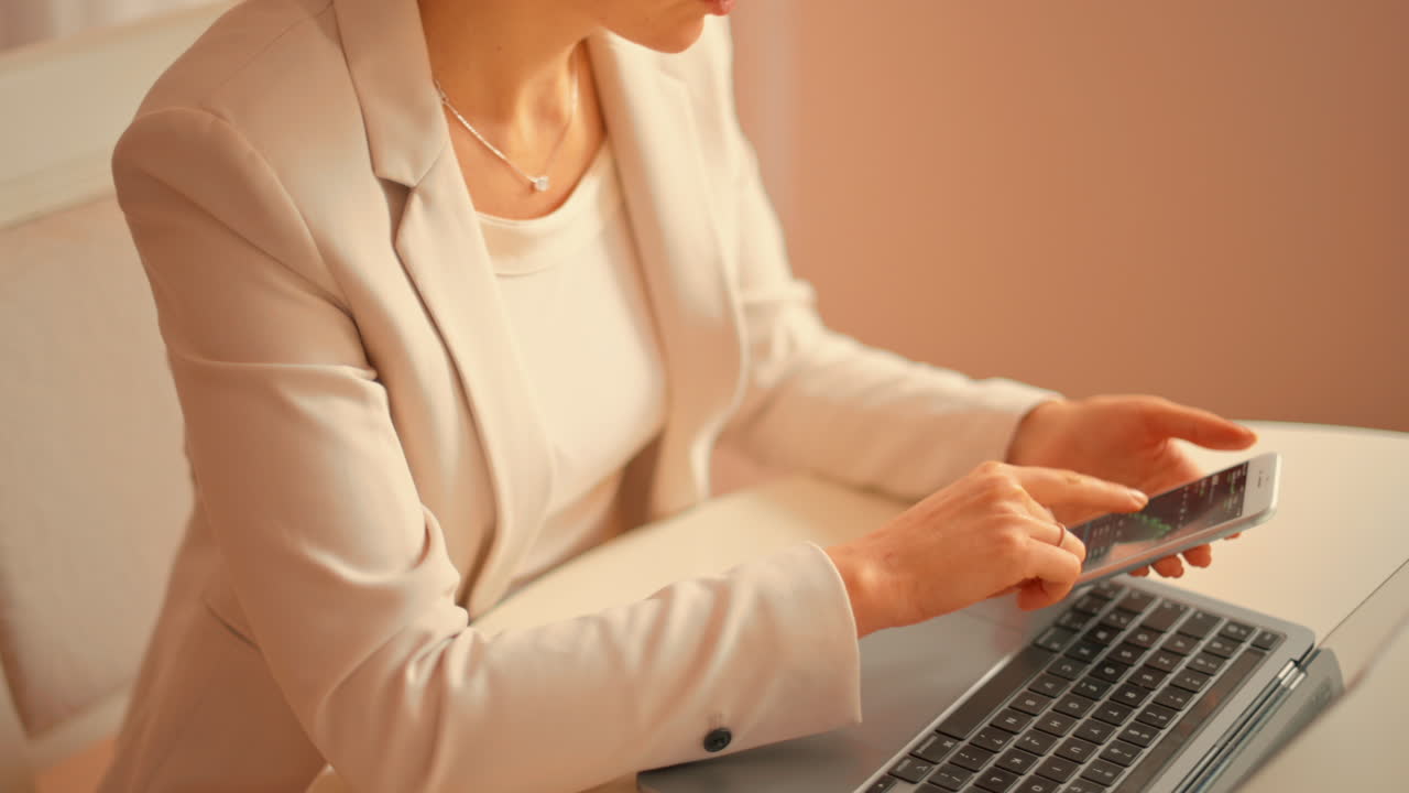 Woman in a white blazer working on a laptop at a table while holding her phone