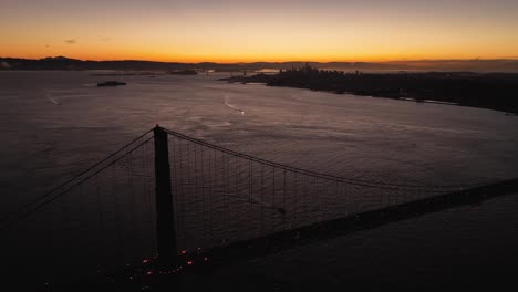 drone view of san francisco bay with silhouette of golden gate bridge in foreground. a beautiful orange sunrise horizon in the distance.