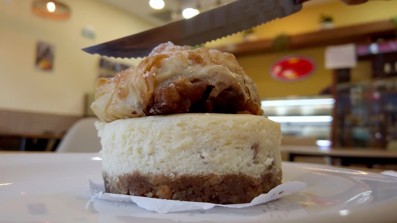 Close up shot of a knife slicing into a rich cheesecake topped with a layer of crispy baklava in a bakery setting