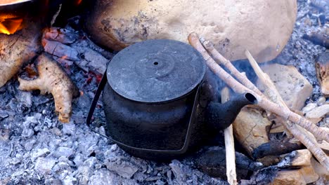 Small-black-teapot-heating-up-in-the-ashes-and-embers-of-open-campfire-in-natural-outdoors-wilderness-environment