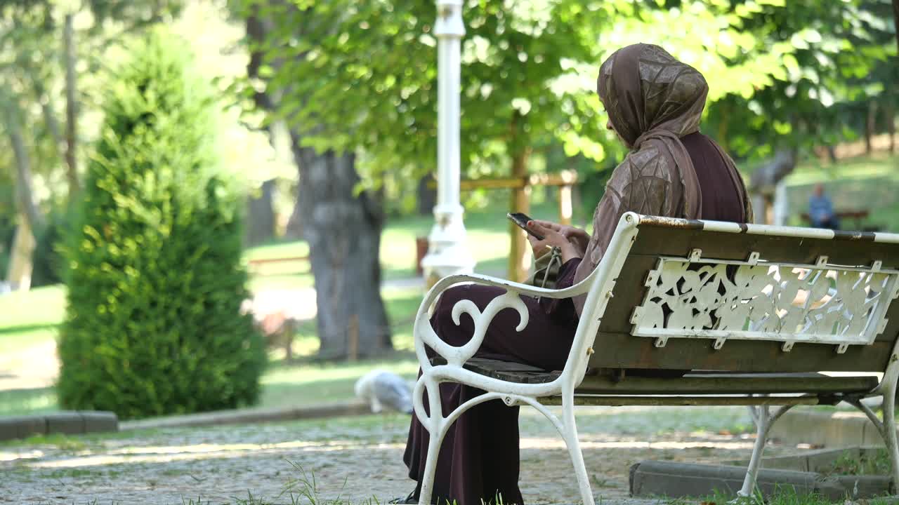 Žena v hidžábu sedí na lavičce v parku a používá svůj telefon