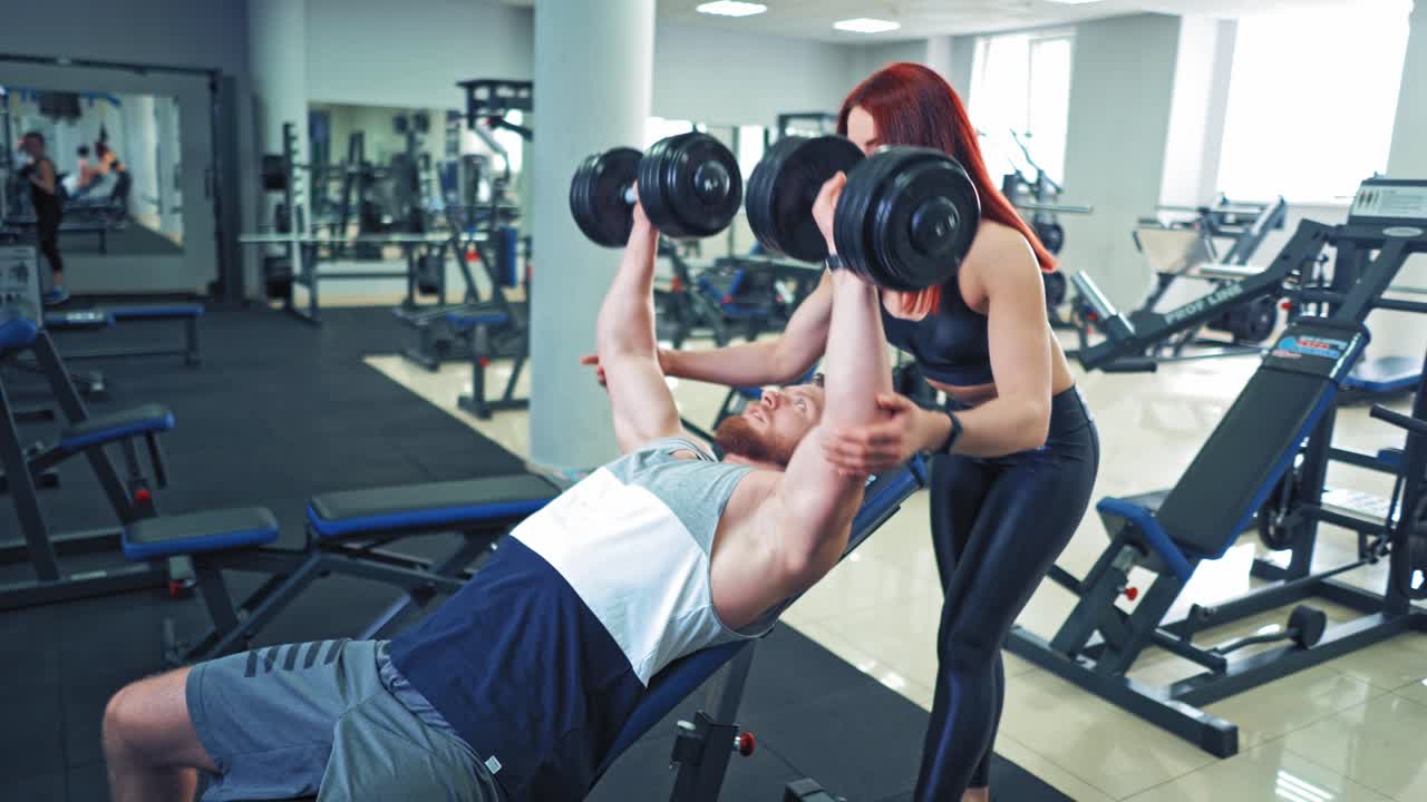 Male athlete is lifting dumbbells while lying on special desk in gym. Bodybuilder doing his daily workout with dumbbells and a woman instructor controls its process.