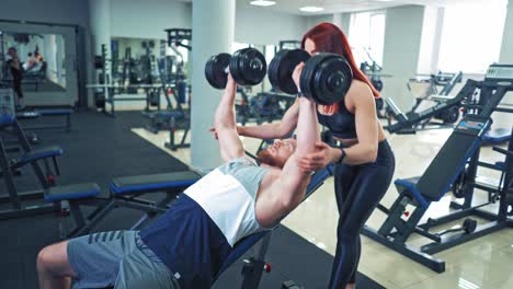 male athlete is lifting dumbbells while lying on special desk in gym. bodybuilder doing his daily workout with dumbbells and a woman instructor controls its process.