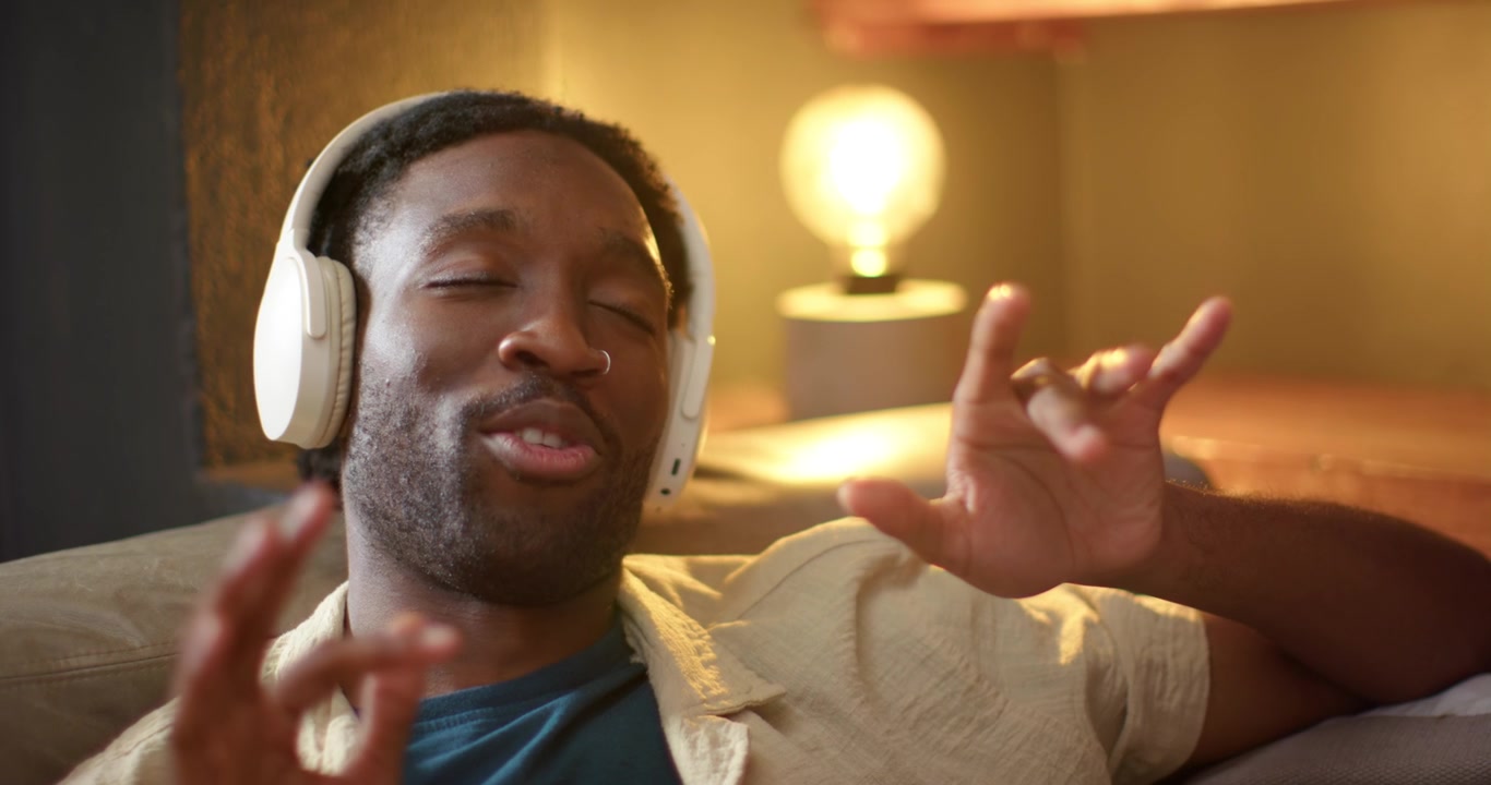Adult African American male reclining on sofa listening and following music with white headphones