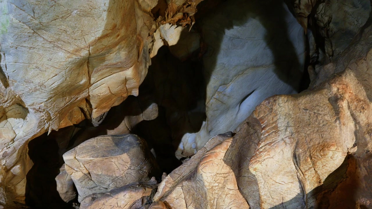 Illuminated limestone rock formations inside a natural cave system reveal layered geological structures and deep shadows. Smooth overhead tracking shot showcases the textured ceiling and walls.