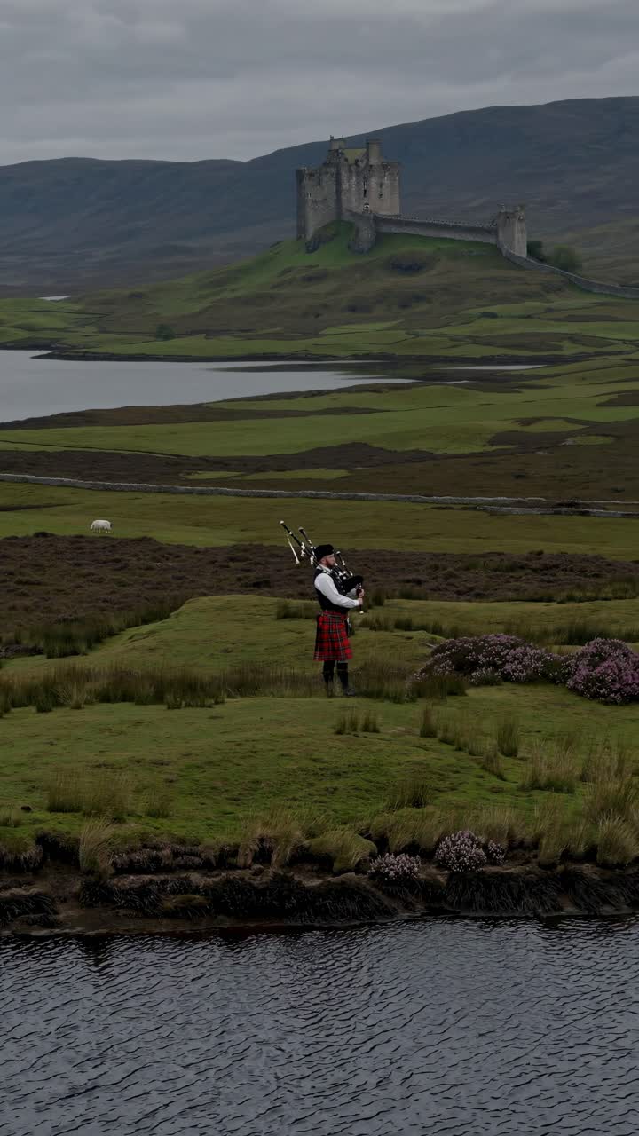 Aerial video of a bagpiper in traditional attire on a grassy hill, with a castle and lake