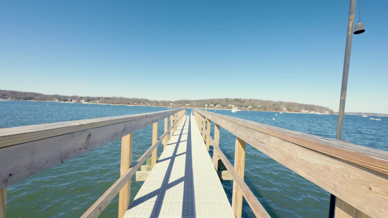 Person walking across a wooden footbridge toward calm coastal waters in Northport, Long Island. Peaceful setting surrounded by trees and bay views on a bright, tranquil day.