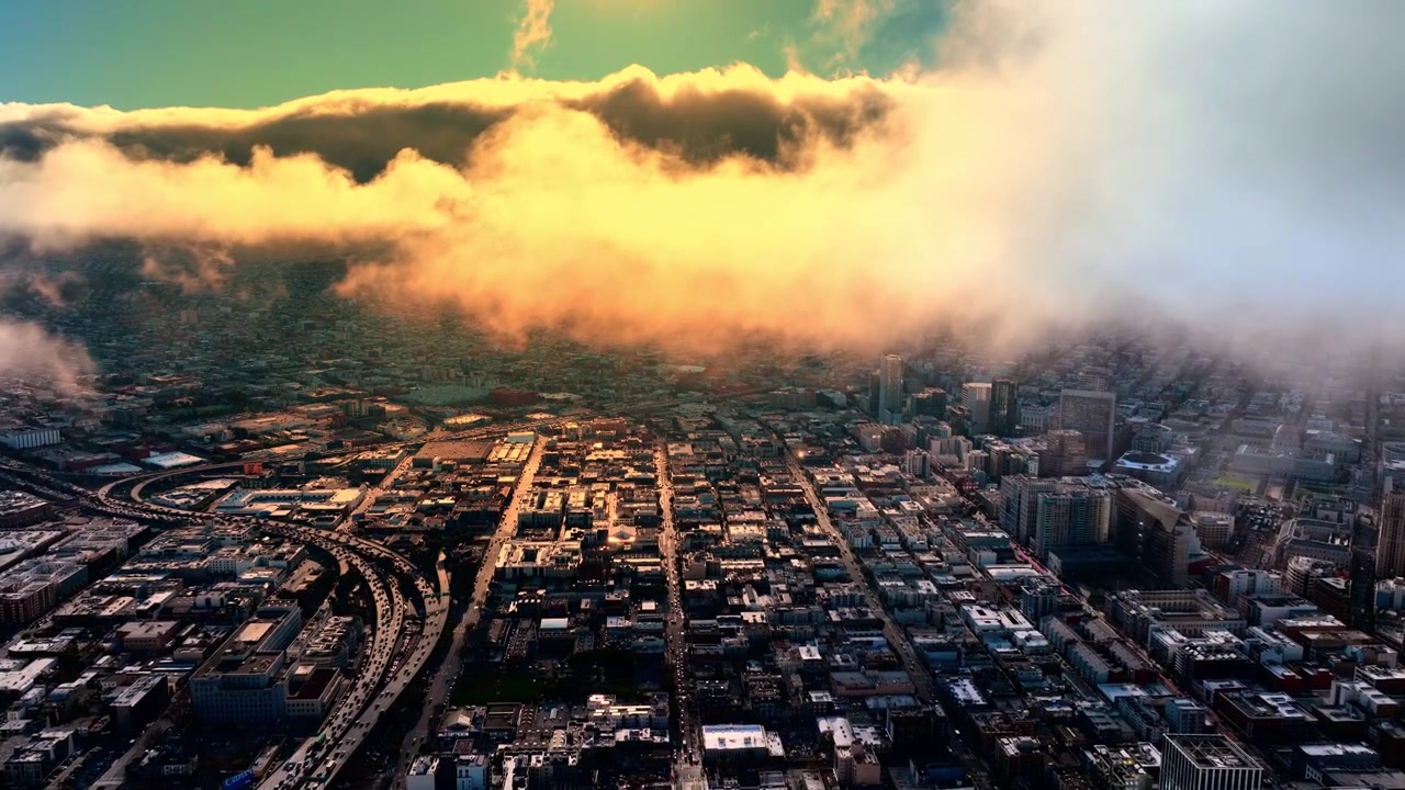 Low cloud layer rolling over urban grid at dusk, San Francisco. Golden lit fog bank descending on flat city blocks, freeway curve at left, teal and amber sky above.