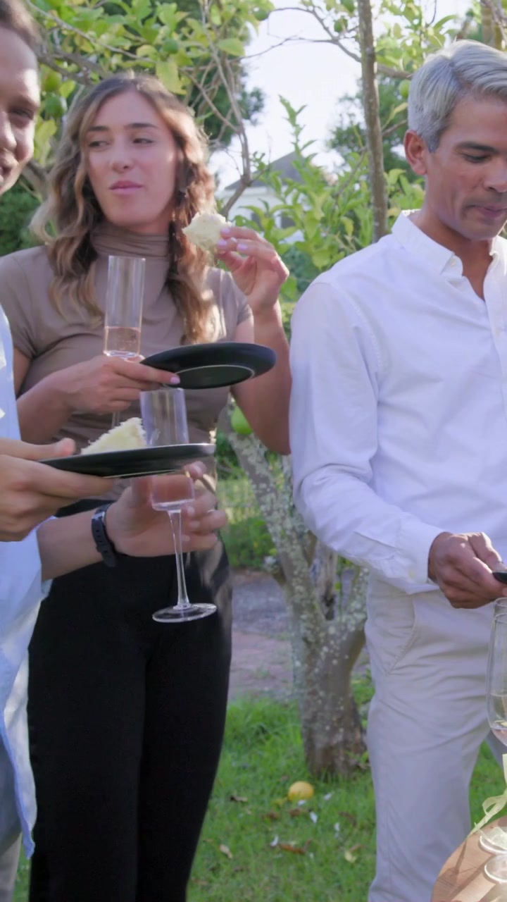 Vídeo vertical: Trio rindo comendo bolo e bebendo em taças de champanhe em um encontro no jardim, com pratos pretos