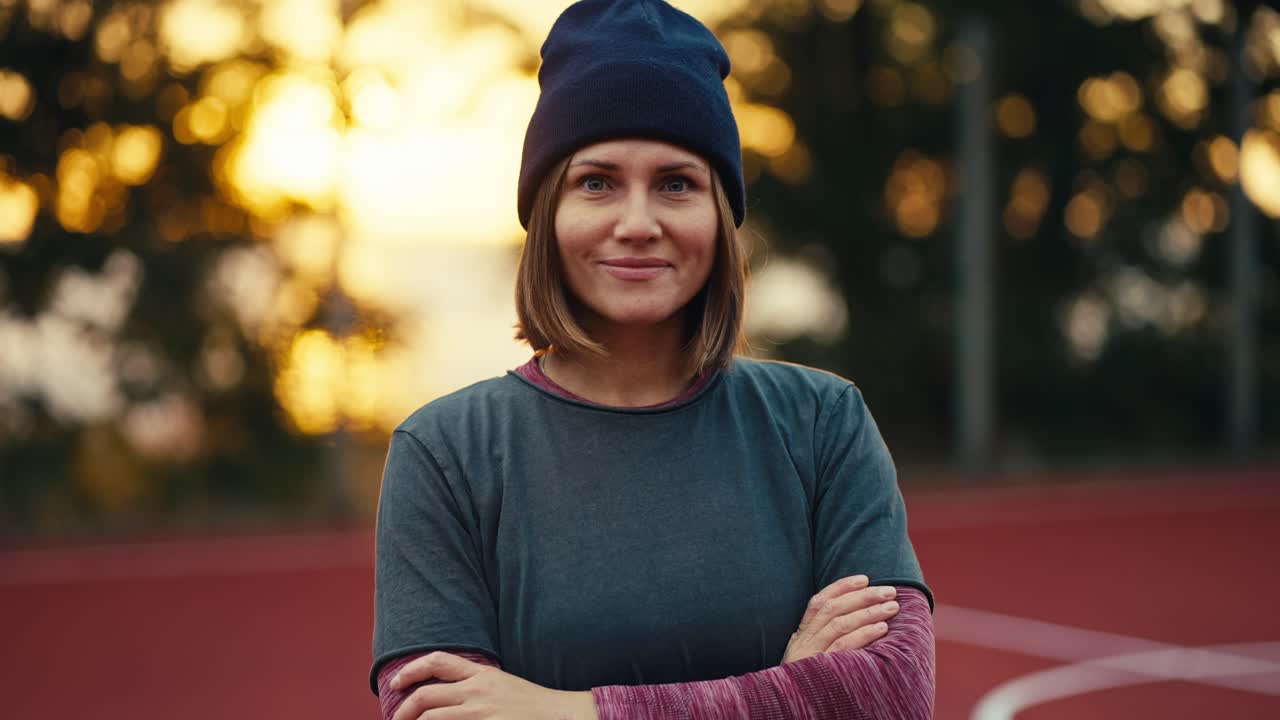 Close-up portrait of a confident and happy blonde girl in a black hat and in a sports uniform who folds her arms on her chest and looks at the camera in the morning at Sunrise