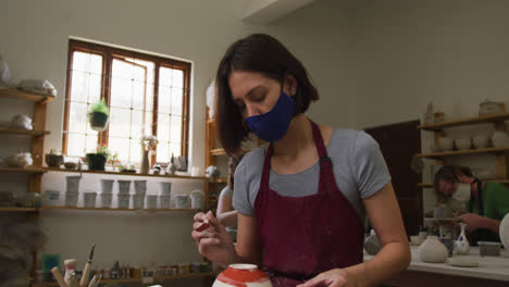 female caucasian potter wearing face mask and apron using glaze brush to paint pot on potters wheel