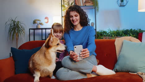 cheerful mother and daughter child girl taking selfie with corgi dog using smartphone on home sofa
