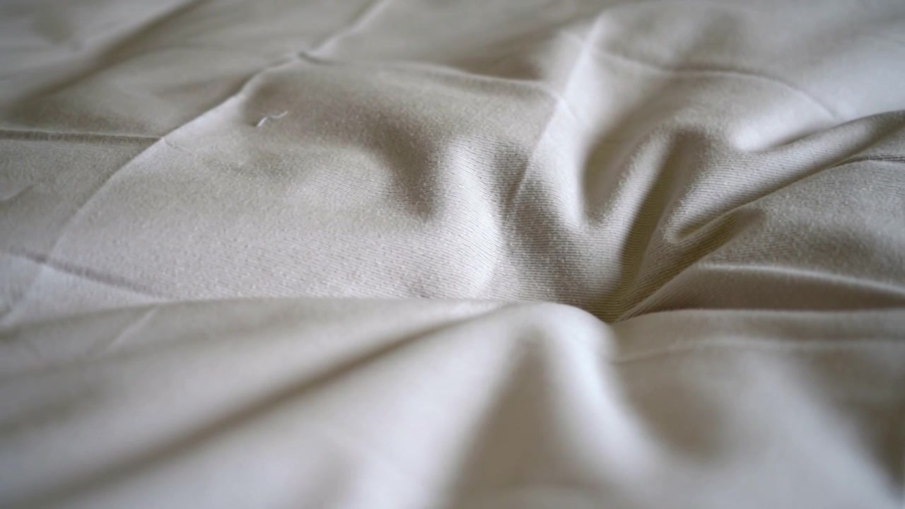 Mostrando un materasso trapuntato bianco sporco che si assesta con sottili cambiamenti di luce in un primo piano della camera da letto, cuciture.