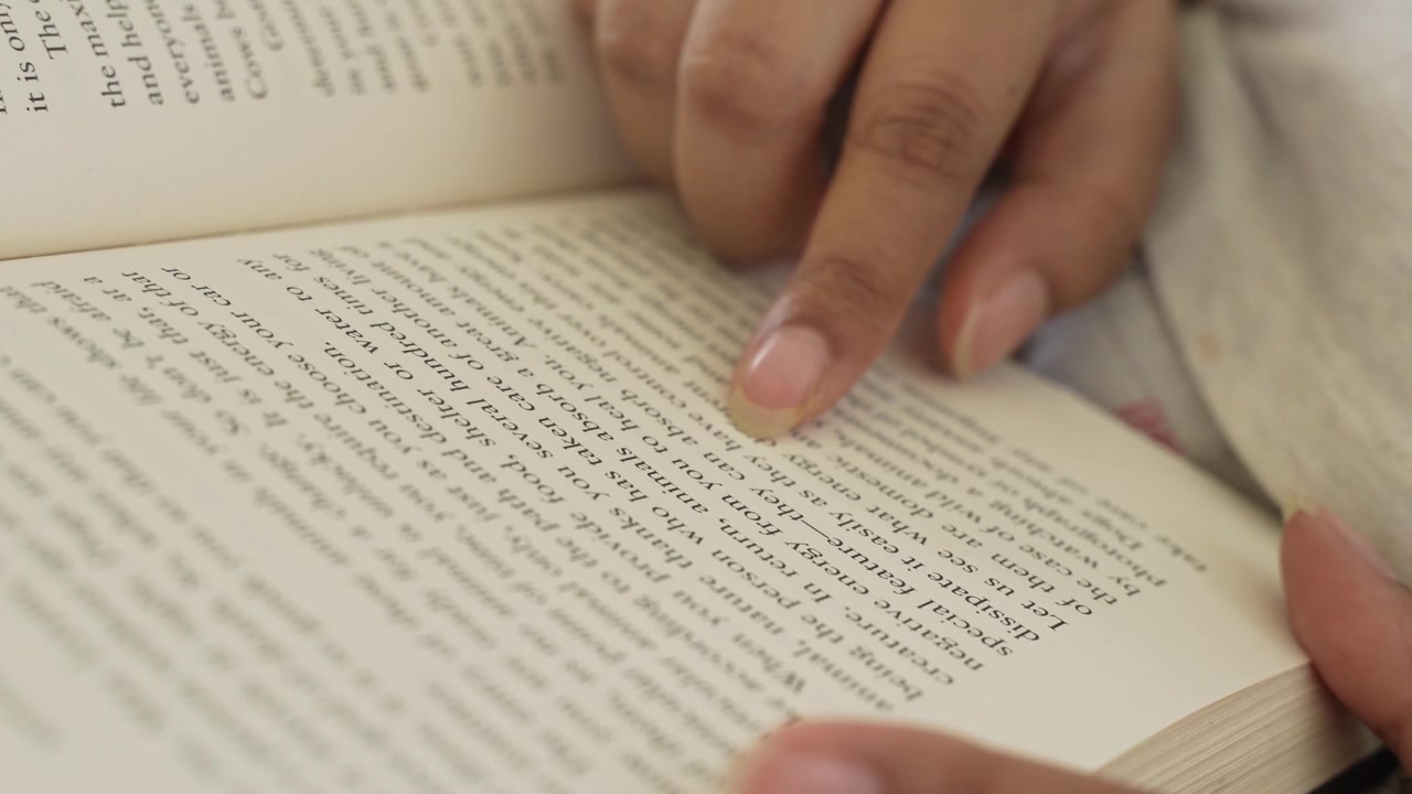 Close-up of a woman's hand gently pressing and smoothing an open book page while reading, with clearly visible printed text and soft natural window light illuminating.