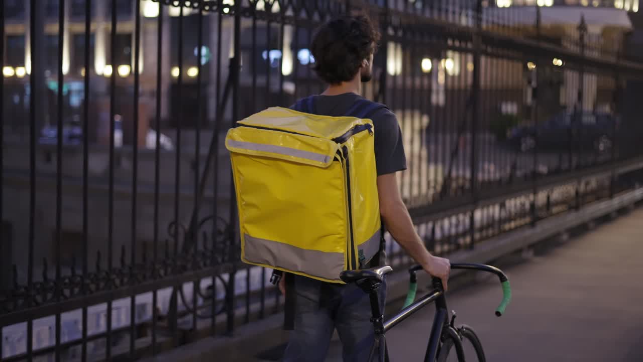 Delivery man walking with bike and yellow bag by city bridge, rear view, slow motion