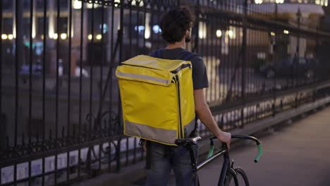 Delivery-man-walking-with-bike-and-yellow-bag-by-city-bridge,-rear-view,-slow-motion