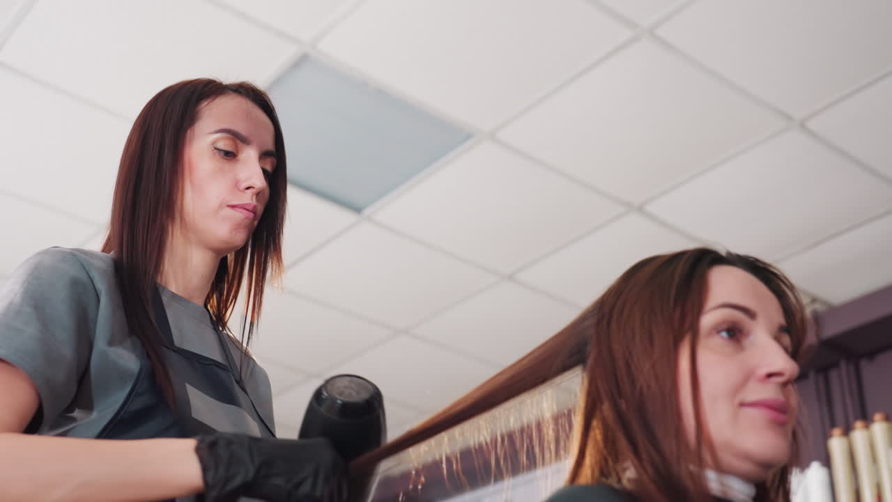 En una peluquería, una estilista caucásica divide el pelo en secciones y lo seca con una técnica muy concentrada; movimiento del secador suspendido, clips bien colocados, manos atentas con guantes, clienta tranquila y sonriente, techo clínico de la peluquería