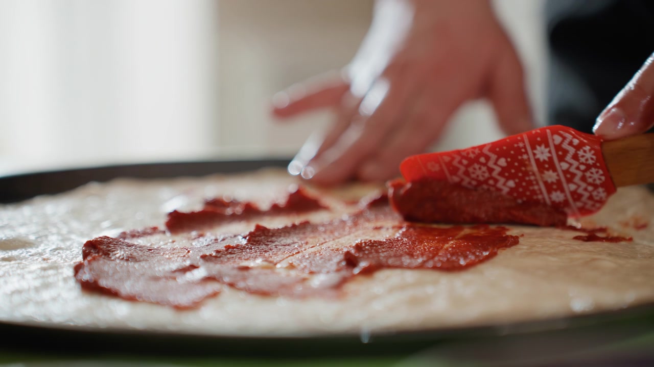 Una vista ravvicinata mostra mani che usano una piccola spatola rossa per stendere la salsa sull'impasto con movimenti costanti, formando strati irregolari, striature, creste, macchie lucide e un'azione vivace in cucina, con mano e salsa.